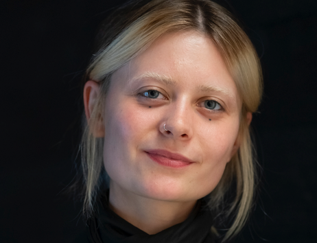 Porträt einer jungen Frau mit blonden Haaren und Nasenpiercing vor schwarzem Hintergrund.