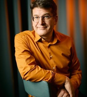 Person in orangefarbenem Hemd sitzt auf einem Stuhl vor einem gestreiften Hintergrund.