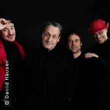 Vier Männer in schwarzer Kleidung vor schwarzem Hintergrund, einer trägt eine rote Mütze.