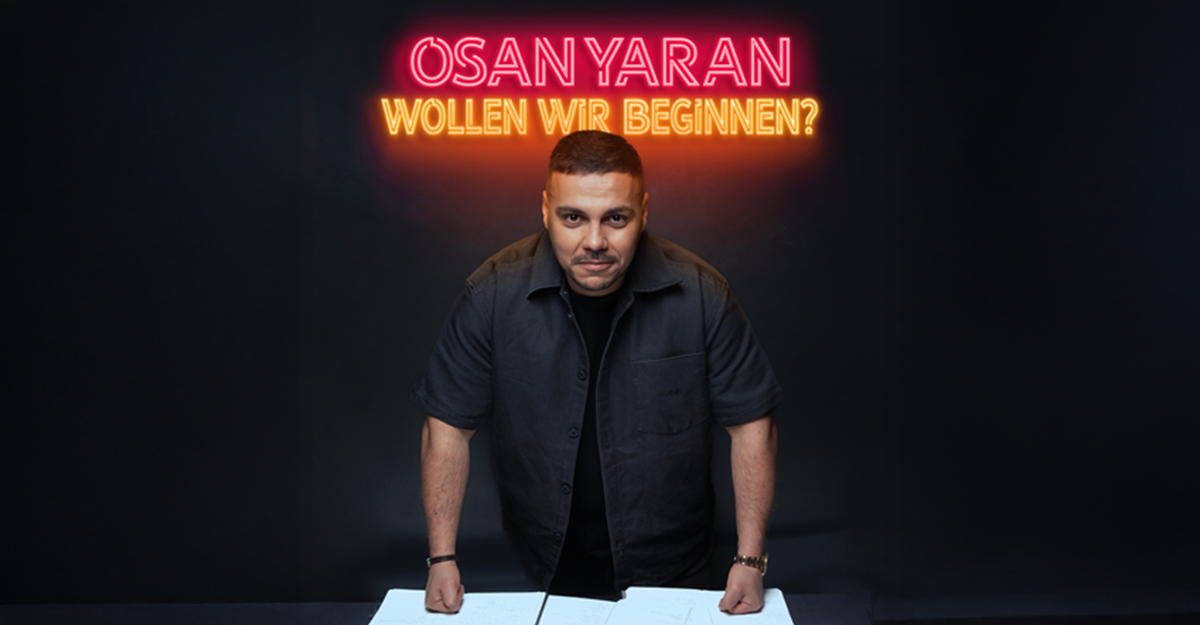 Ein Mann steht vor einem Tisch mit Neon-Schriftzug 'Osan Yaran - Wollen wir beginnen?' im Hintergrund.