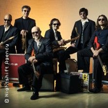 Eine Bluesband mit sechs Mitgliedern, die Instrumente wie Saxophon und Gitarre halten, posiert vor einem neutralen Hintergrund.