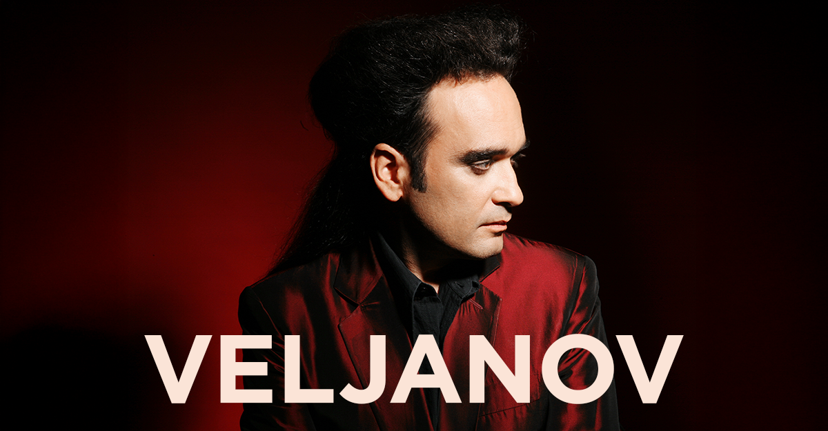 Person in rotem Anzug vor rotem Hintergrund mit dem Schriftzug 'VELJANOV'.