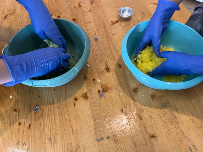Zwei Personen mit blauen Handschuhen mischen bunte Zutaten in Schüsseln auf einem Holztisch.