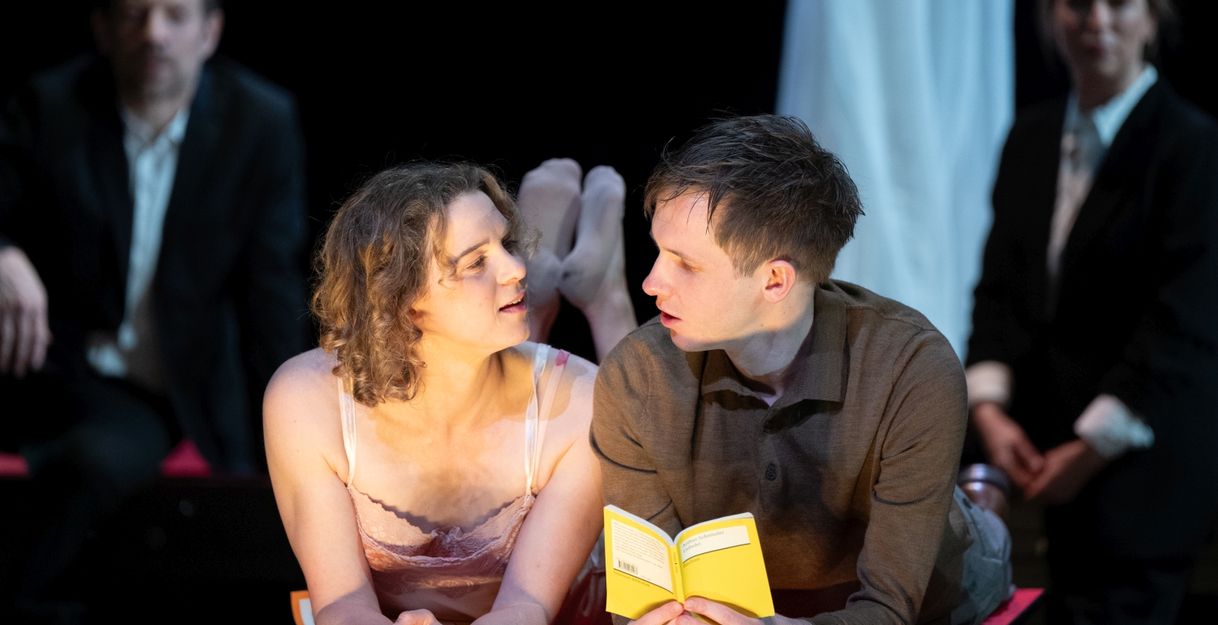 Zwei Personen liegen auf einem Tisch und schauen sich an, während eine Person ein Buch hält. Im Hintergrund sitzen zwei weitere Personen.