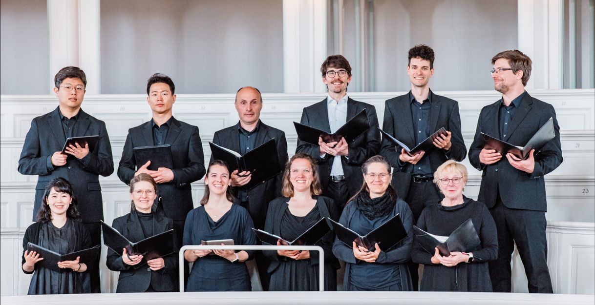 Eine Gruppe von zwölf Chorsängern in schwarzer Kleidung steht in einem Kirchenraum und hält Notenblätter.