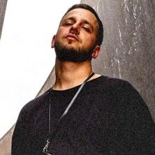 Ein Mann mit Bart und schwarzem Oberteil steht vor einer Betonwand und blickt selbstbewusst in die Kamera.