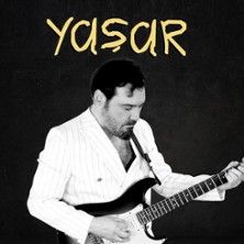 Ein Musiker in einem weißen Anzug spielt E-Gitarre vor einem dunklen Hintergrund. Oben steht der Name "Yasar".