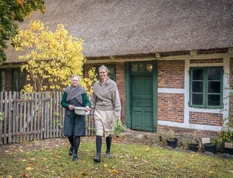 Zwei Frauen in historischer Kleidung vor einem reetgedeckten Fachwerkhaus mit grünem Türrahmen und herbstlichem Garten.