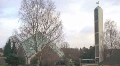 Kirche mit modernem Turm und dreieckigem Dach, umgeben von Bäumen.