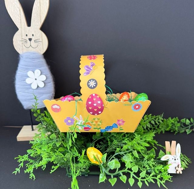 Ein dekoratives Osternest mit bunten Eiern, umgeben von grünen Pflanzen. Daneben steht ein hölzerner Osterhase.