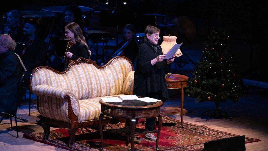 Eine Frau liest auf einer Bühne vor einem Sofa, mit einem Weihnachtsbaum im Hintergrund.