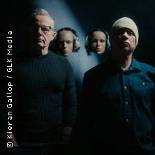 Zwei Männer stehen in einem dunklen Raum, im Hintergrund sind zwei Masken mit Kopfhörern zu sehen.