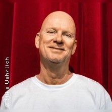 Ein lächelnder Mann in einem weißen T-Shirt steht vor einem roten Vorhang.