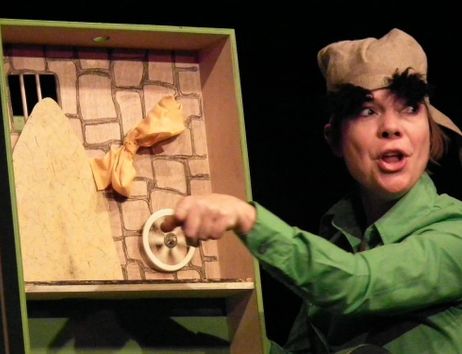 Eine Person in einem grünen Kostüm spielt in einem Puppentheater. Sie trägt eine Mütze und zeigt auf ein hölzernes Bühnenbild mit einem Rad.