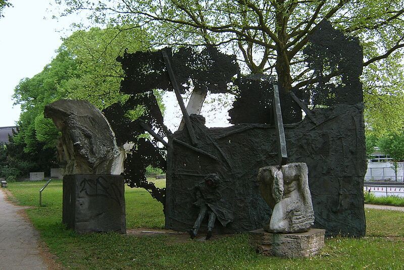 Foto eines Denkmals für den 2. Weltkrieg, das aus drei Teilen besteht. Zwei kleinere abstrakte Elemente aus Stein stehen je auf einem Steinsockel. Das größte Element ist schwarz, flächig und eine Person ragt mit Beinen und Mantel heraus, ebenso Stangen.