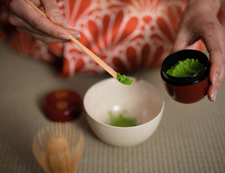 Matcha wird mit einem Bambuslöffel in die Schale gefüllt. Die Frau trägt einen roten Kimono mit weißem Muster drauf.