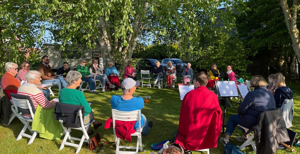 Eine Gruppe von Menschen sitzt im Kreis auf weißen Stühlen im Freien unter Bäumen.
