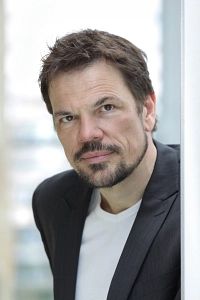 Porträt eines Mannes mit Bart in einem schwarzen Sakko und weißem T-Shirt.