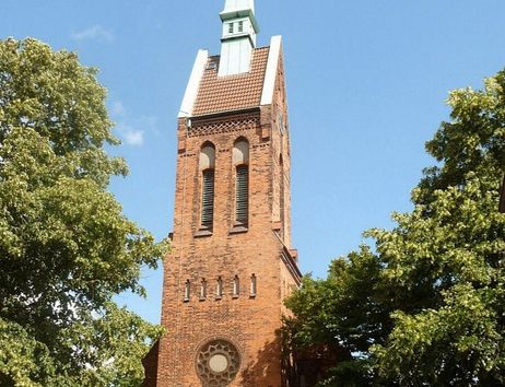 Backsteinkirche mit hohem Turm und grünem Spitzdach, umgeben von Bäumen.