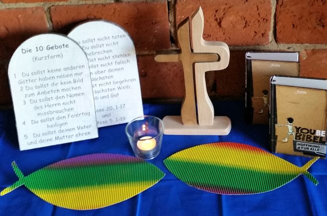 Religiöse Tischdekoration mit Kreuz, Bibeln, Kerze und den 10 Geboten vor einer Backsteinwand.
