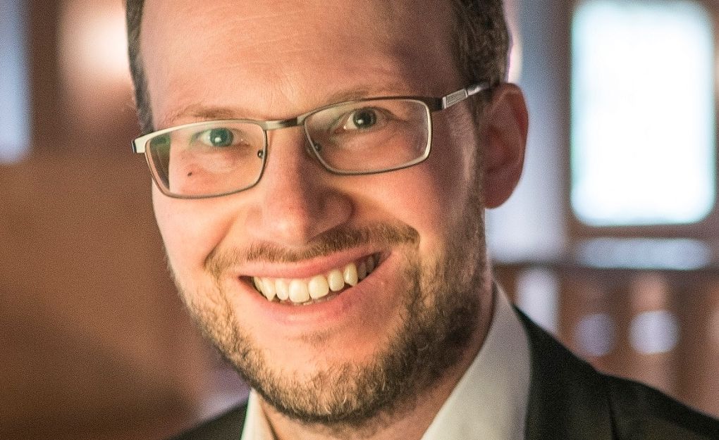 Porträt eines lächelnden Mannes mit Brille und Bart in einem Anzug.