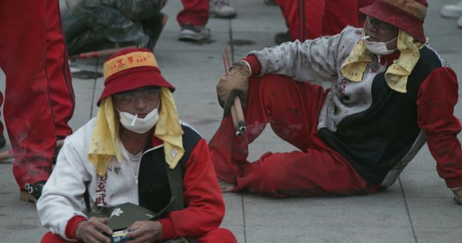 Sie sehen ein Filmstill aus Yalda Afsahs Arbeit HEAT: Zwei Männer sitzen auf einem Steinboden. Sie tragen rot-weiße Kleidung, Hüte mit taiwanischem Schriftzug, Schutzbrillen und Mundschutz. Beide rauchen, einer hält Feuerwerkskörper in der Hand.