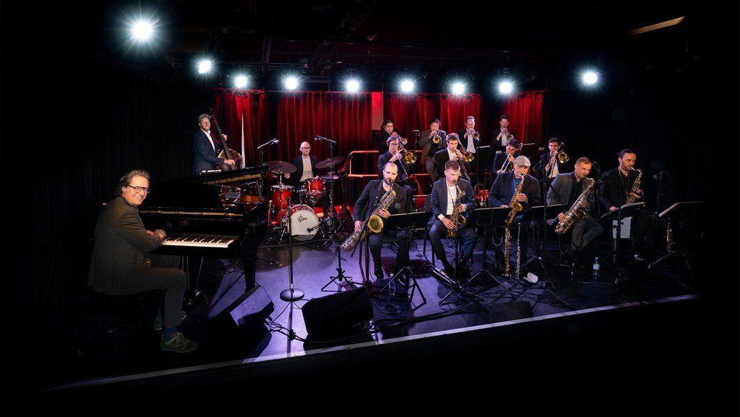 Eine Jazz-Big-Band auf einer Bühne mit rotem Vorhang und Beleuchtung. Ein Pianist sitzt links, während andere Musiker Blasinstrumente spielen.