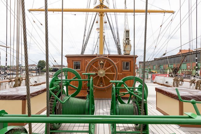 Deck eines historischen Segelschiffs mit Steuerrad und grüner Winde.