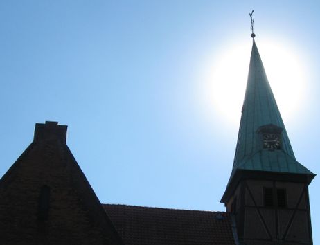 Kirchturm mit Uhr vor strahlend blauem Himmel und Sonne im Hintergrund.