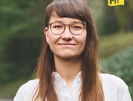 Porträt einer Frau mit Brille und langen Haaren, die ein weißes Hemd trägt. Im Hintergrund ist verschwommenes Grün zu sehen. Oben rechts ist ein gelbes Logo mit den Buchstaben 'HF'.