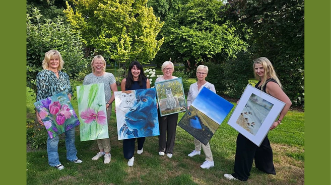 Sechs Frauen der Malgruppe aus Glinstedt stehen im Garten und präsentieren ihre gerahmten Bilder bei der Kunstausstellung.