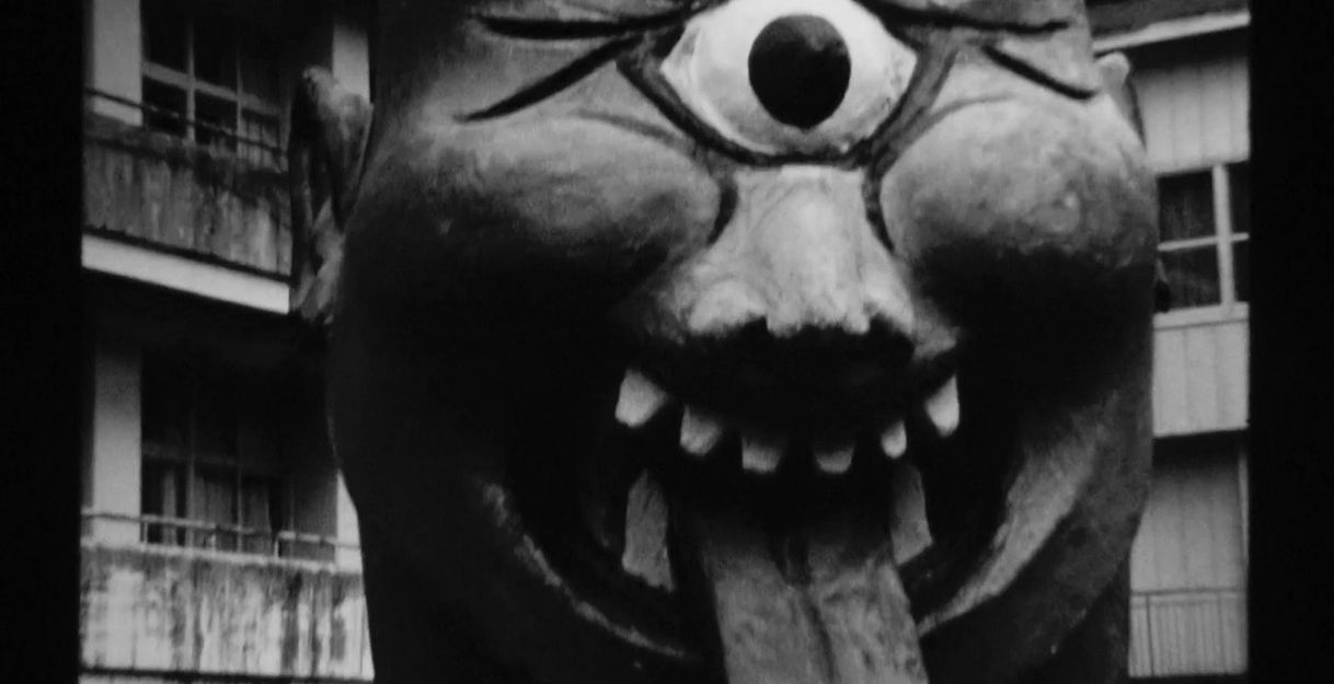 Schwarz-weißes Bild einer grotesken Skulptur mit einem Auge und herausgestreckter Zunge vor einem Gebäude.