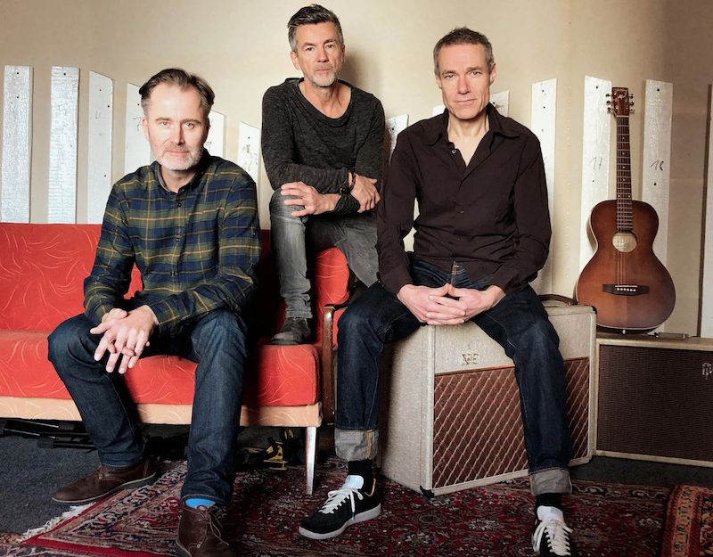 Drei Männer sitzen in einem Raum mit einer Gitarre im Hintergrund.