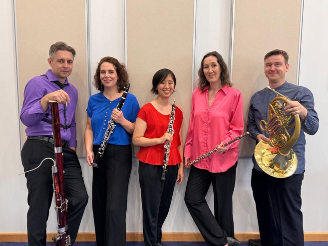 Fünf Musiker mit Holzblasinstrumenten und einem Horn posieren vor einer Wand.