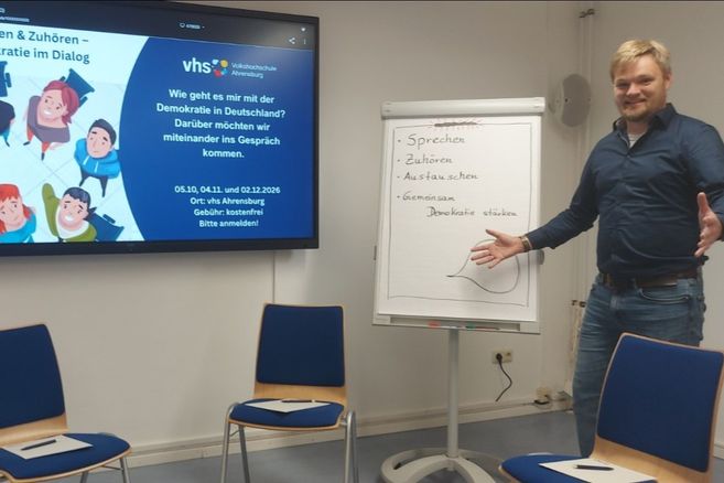 Mann steht in einem Seminarraum neben einem Flipchart und einem Bildschirm mit der VHS Ahrensburg zu Demokratie in Deutschland; leere Stühle im Vordergrund.