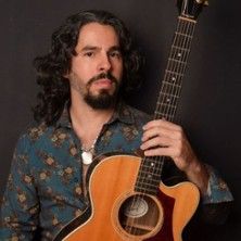 Ein Mann mit langen Haaren hält eine Akustikgitarre vor einem dunklen Hintergrund.