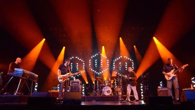 Eine Band spielt auf einer Bühne mit orangefarbenem Licht und Lichteffekten im Hintergrund.
