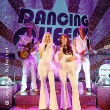 Eine Band in weißen Outfits performt auf einer Bühne mit dem Schriftzug 'Dancing Queens'. Zwei Sängerinnen stehen vorne, begleitet von zwei Musikern im Hintergrund.