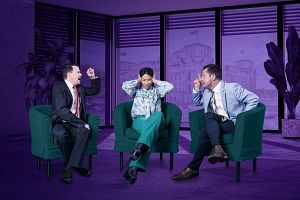 Drei Personen sitzen in grünen Sesseln in einem lila Raum. Zwei Personen gestikulieren, während die dritte Person die Ohren zuhält.