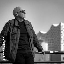 Eine Person mit Sonnenbrille lehnt an einem Geländer, im Hintergrund ist ein modernes Gebäude zu sehen.