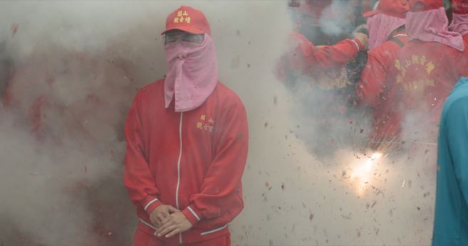 Sie sehen ein Filmstill aus der Arbeit HEAT von Yalda Afsah. Im Mittelpunkt ist das Porträt eines Teilnehmers der taiwanischen Feuerwerkszeremonie zu sehen. Die Person trägt eine Brille und hat ein Tuch vor Mund und Nase. Dichter Rauch umgibt ihren Kopf und Oberkörper.