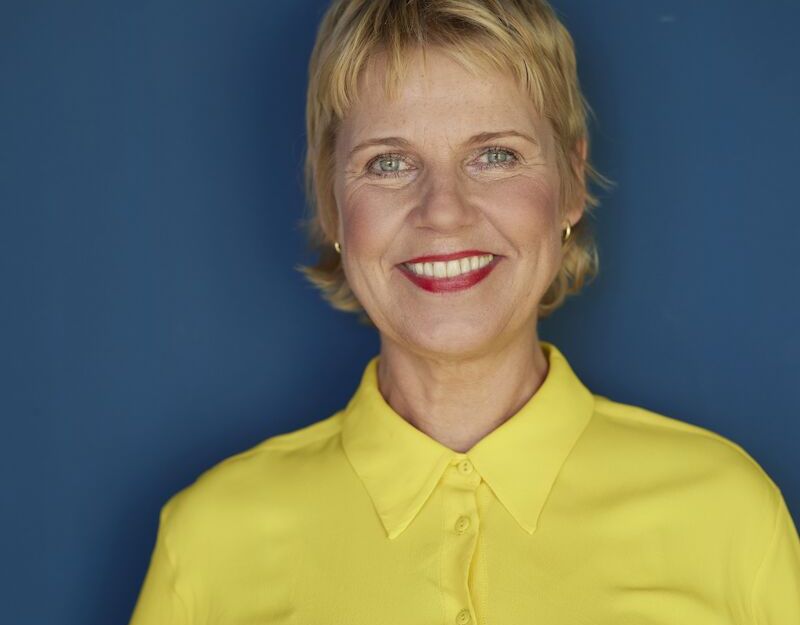 Frau in gelber Bluse vor blauem Hintergrund lächelt in die Kamera.