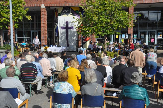 Menschen versammeln sich zu einem Open-Air-Gottesdienst vor einem Gebäude mit einem großen Kreuz auf einem Tuch.