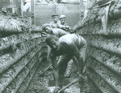 Arbeiter graben in einem schmalen, erdigen Graben mit Schaufeln.