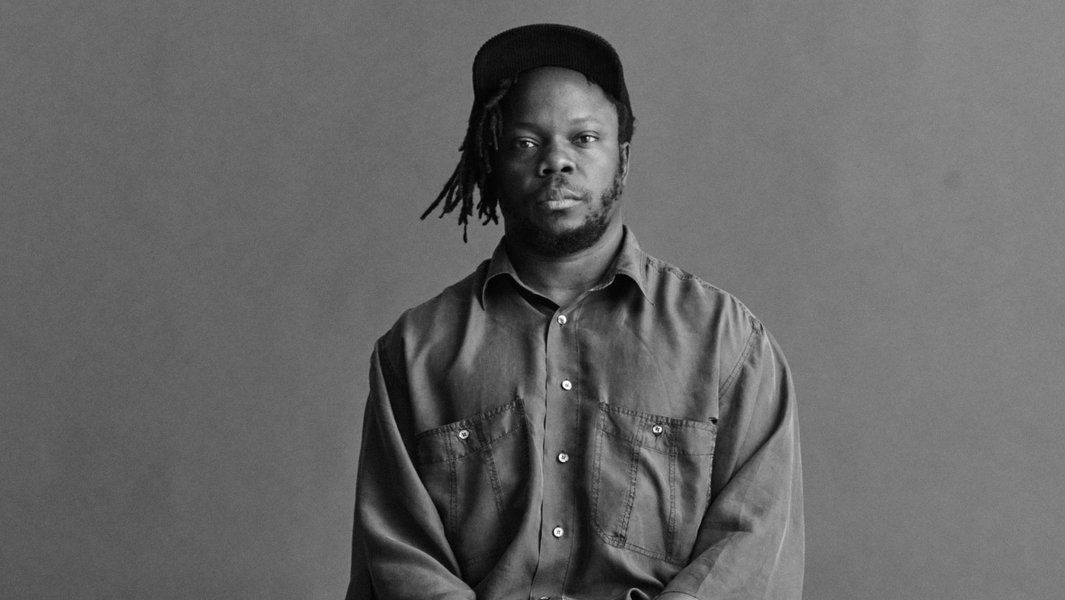 Schwarz-weiß Porträt eines Mannes mit Dreadlocks und Basecap, der in die Kamera schaut.