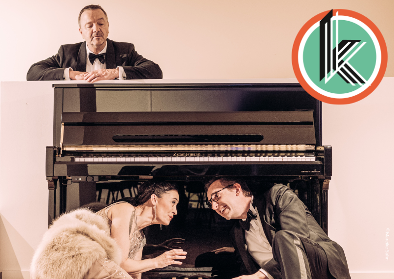 Drei Personen in Abendkleidung posieren um ein Klavier. Zwei sitzen darunter, eine lehnt sich oben darauf. Ein Logo ist in der oberen rechten Ecke.